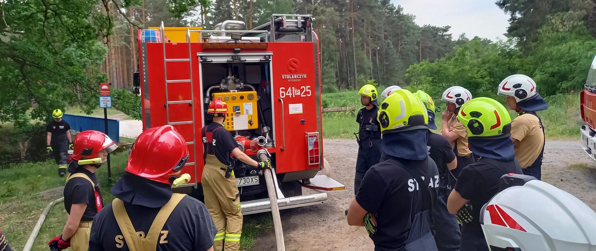 Strażacy stoją zgromadzeni przy samochodzie gaśniczym. Instruktor obsługuje autopompę. W tle widać las.