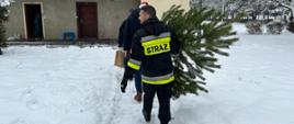 Strażak, Policjant i pracownik MOPS idą jeden za drugim w kierunku budynku mieszkalnego z choinką, ozdobami świątecznymi. Na plecach ostatniego z nich napis STRAŻ. Na ziemi leży śnieg. W prawej części zdjęcia ośnieżone drzewo.