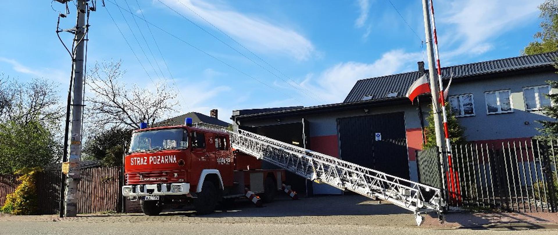 Samochód pożarniczy z wysunięta w bok i do dołu mechaniczną drabiną pożarniczą stoi przed budynkiem strażnicy OSP Boża Wola