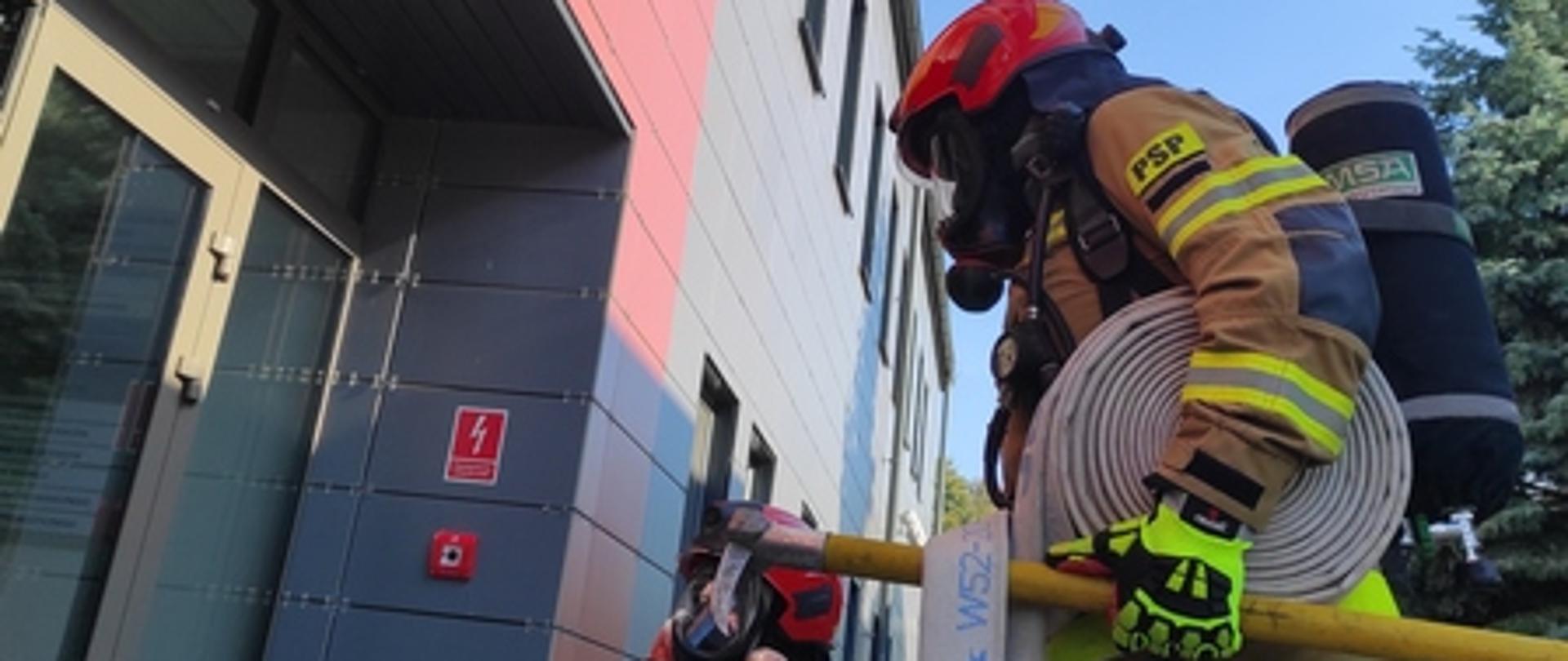 Strażacy w mundurach bojowych przed wejściem do budynku. Z okna budynku wydobywa się dym.