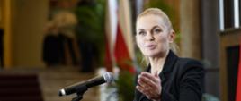 Zdjęcie z boku, minister Nowacka stoi i mówi do mikrofonu na stojaku, za nią w tle widok na korytarz i nakryte fioletowym dywanem schody.