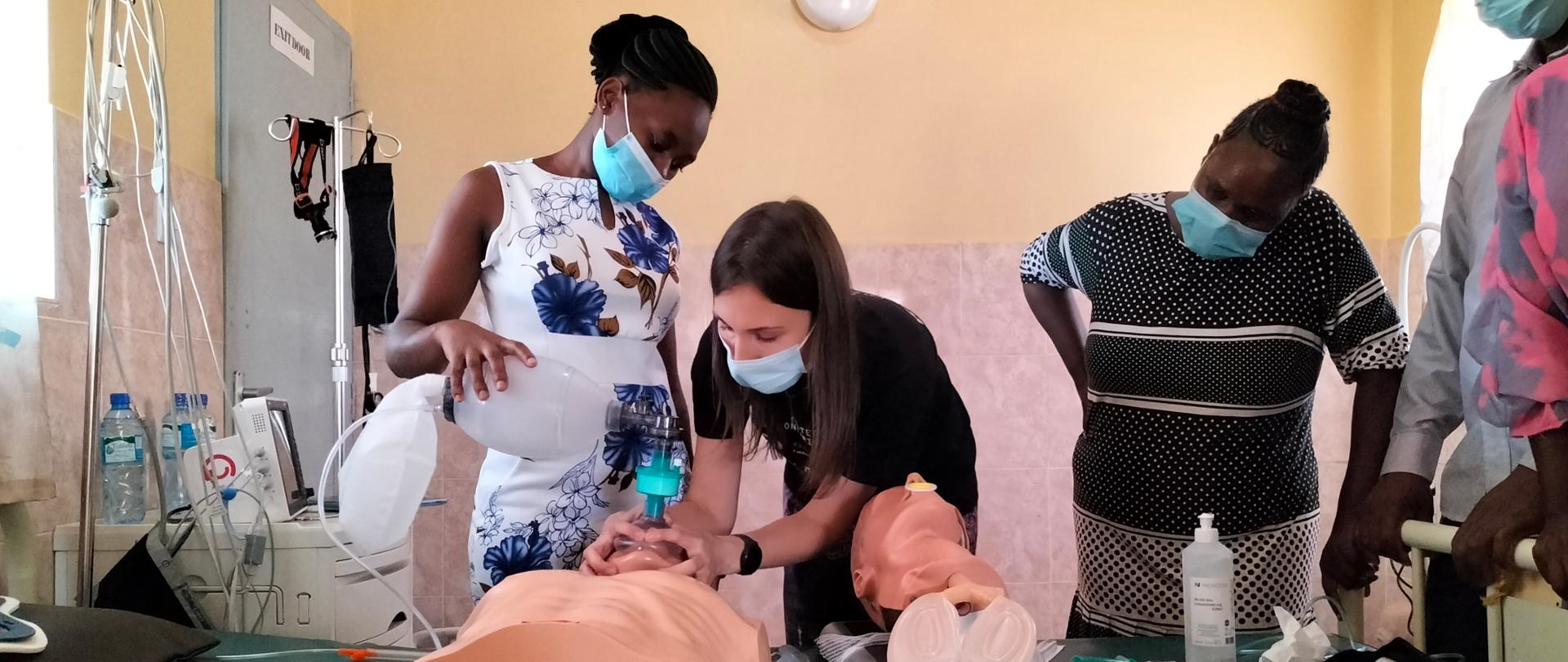 Pięć osób w maseczkach znajduje się w sali medycznej, pochylają się nad fantomami. Dwie kobiety intubują fantoma.