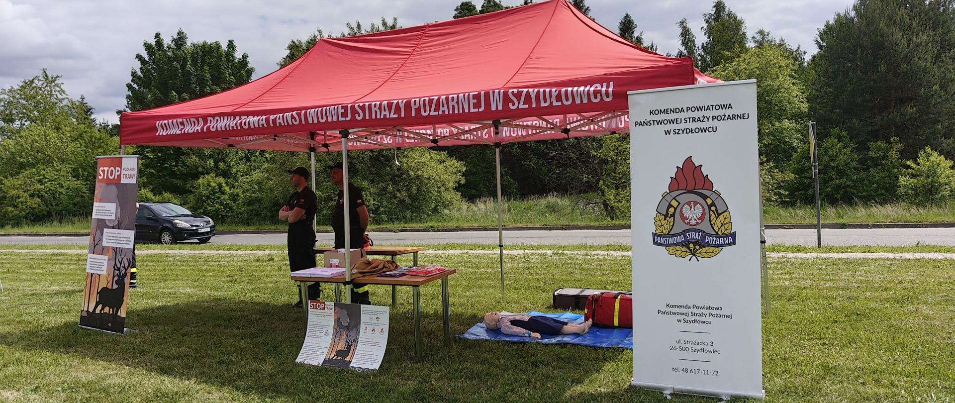 Zdjęcie przedstawia plenerowe stoisko edukacyjno-promocyjne przygotowane przez strażaków. Stanowisko zostało rozstawione na zielonej trawie w pobliżu drogi, w otoczeniu drzew. Służy ono celom instruktażowym, m.in. nauce pierwszej pomocy oraz profilaktyce przeciwpożarowej.