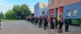 Strażacy stoją trójkami i trzymają poczty sztandarowe.