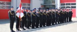 Na tle budynku garażowego pododdział strażaków stojący w umundurowaniu galowym w dwuszeregu. Z lewej strony Sztandar Komendy Powiatowej Państwowej Straży Pożarnej w Lublińcu.