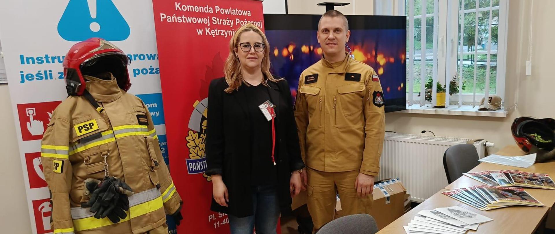 Na zdjęciu jest widocznych dwóch pracowników (kobieta i mężczyzna) Komendy Powiatowej PSP w Kętrzynie (KP PSP). Za nimi widoczny baner KP PSP w Kętrzynie, monitor oraz ubranie specjalne strażaka