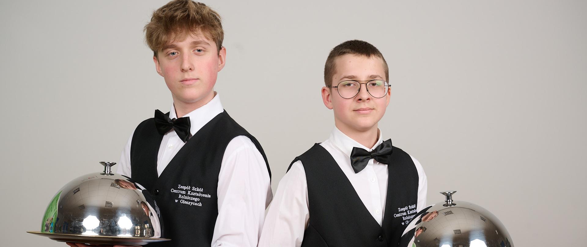 Dwóch uczniów stoi w strojach kelnerskich. Każdy w lewej ręce trzyma tacę do serwowania posiłków oraz przewieszoną serwetę. 