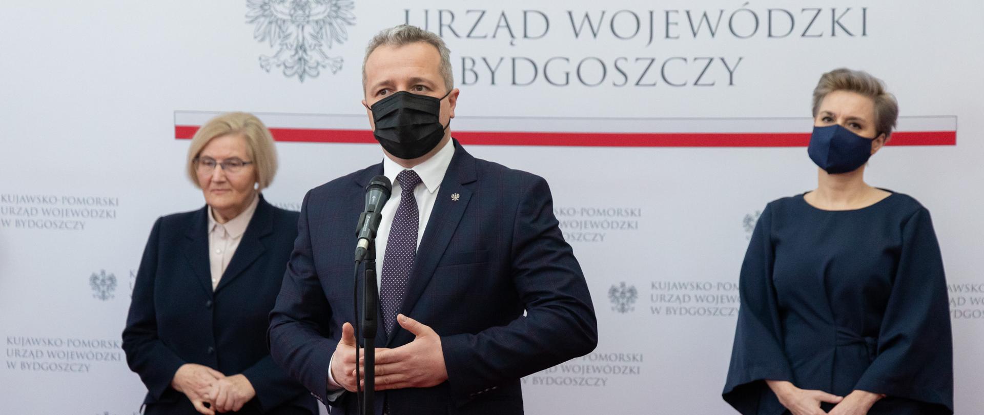Jeden mężczyzna i dwie kobiety stojący na białym tle, mężczyzna wypowiada się do mikrofonu