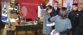 Targi edukacyjne. Sala sportowa w ZSZ w Rawiczu. Widok na stoisko Komendy Powiatowej PSP w Rawiczu. W tle ścianka reklamowa, przed nią na stole znajduje się symulator zagrożeń pożarowych - budynek mieszkalny, a także maskotki strażackie i sprzęt. Przed stołem, z lewej strony ustawiony jest manekin mający na sobie hełm oraz ubranie specjalne. Z prawej strony trzech strażaków udziela informacji uczennicom.