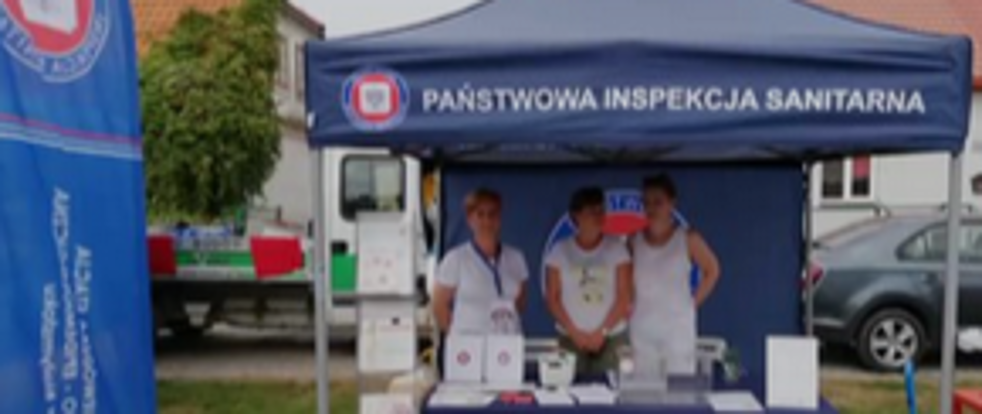 Fotografia plenerowa stoiska PSSE Wysokie Mazowieckie w Tykocinie na XXIV Biesiadzie Miodowej. W centrum pracownicy urzędu z emblematami.