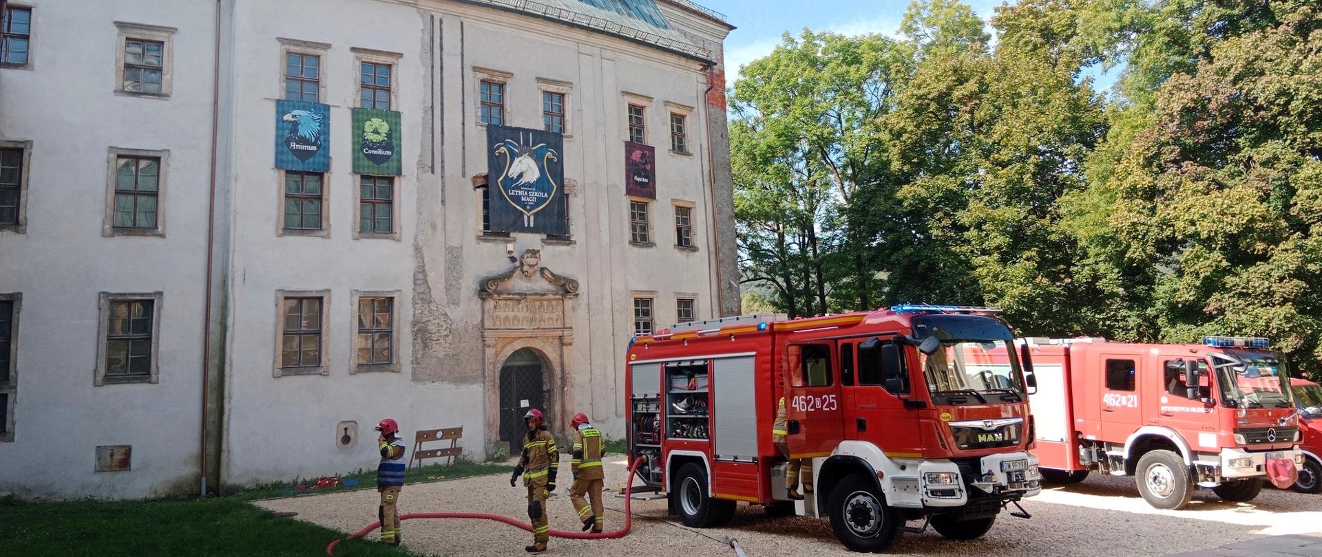 Po lewej stronie planu widok na zamek. Przed zamkiem na placu stoją trzy samochody pożarnicze. Przy pierwszym samochodzie stoi trzech strażaków w umundurowaniu specjalnym przygotowywując się się do ćwiczeń.