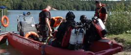 przy brzegu łódź motorowa, w łodzi siedzi dwóch płetwonurków z aparatami powietrznymi na plecach i maską oddechową na twarzy, w łodzi trzech ratowników, maja kapoki na sobie barwy pomarańczowej, ubrani są w koszulki z krótkim rękawem, obok łodzi po prawej stronie stoi strażak z czapka z daszkiem na głowie, ma kamizelkę ratunkową na sobie. Łódź barwy czerwonej z napisem państwowa straż pożarna z numerem bocznym 531EŁ02, w tle rozległy zbiornik wodny, dookoła niego las, przy brzegu zarośla i wysoka trawa, słoneczna pogoda