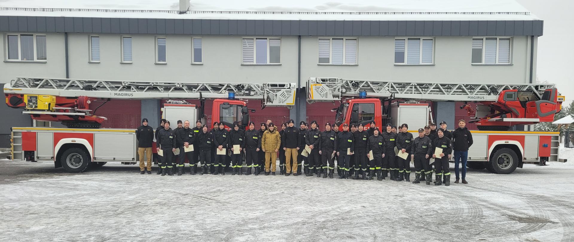 Na zdjęciu widzimy grupę strażaków pozującą do zdjęcia na tle samochodów z drabiną mechaniczną. Strażacy w rękach trzymają certyfikaty potwierdzające udział w szkoleniu z obsługi drabin. 