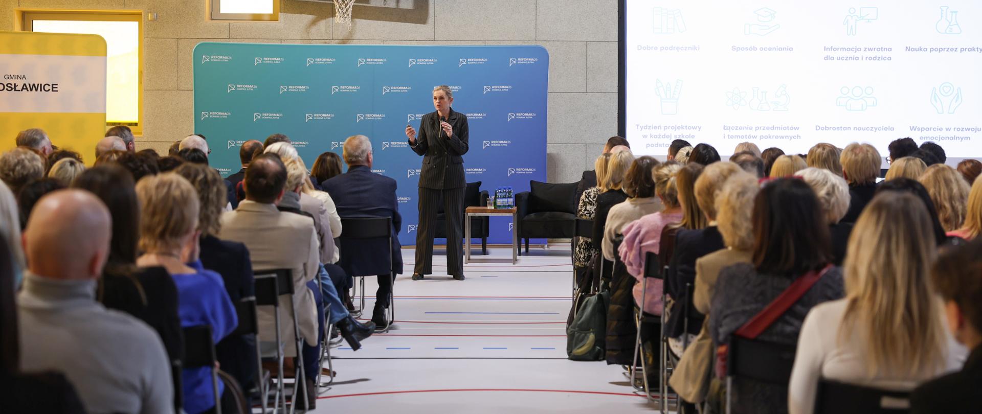 Barbara Nowacka stoi na scenie na tle niebieskiej ścianki brandingowej „REFORMA26”. Po jej prawej stronie znajduje się duży ekran z prezentacją zatytułowaną „NAJWAŻNIEJSZE ZMIANY”, wyświetlający ikony dotyczące m.in. dobrych podręczników i dobrostanu nauczyciela. Na pierwszym planie widoczne są plecy słuchaczy siedzących w rzędach.