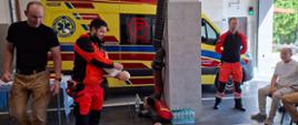 W garażu straży pożarnej odbywa się pokaz pierwszej pomocy. Ratownik medyczny demonstruje czynności ratunkowe z wykorzystaniem fantomu niemowlęcia, a na podłodze leży fantom osoby dorosłej do ćwiczeń resuscytacji. W tle widoczna jest karetka pogotowia oraz strażacy uczestniczący w szkoleniu. Po prawej stronie siedzą słuchacze obserwujący prezentację.