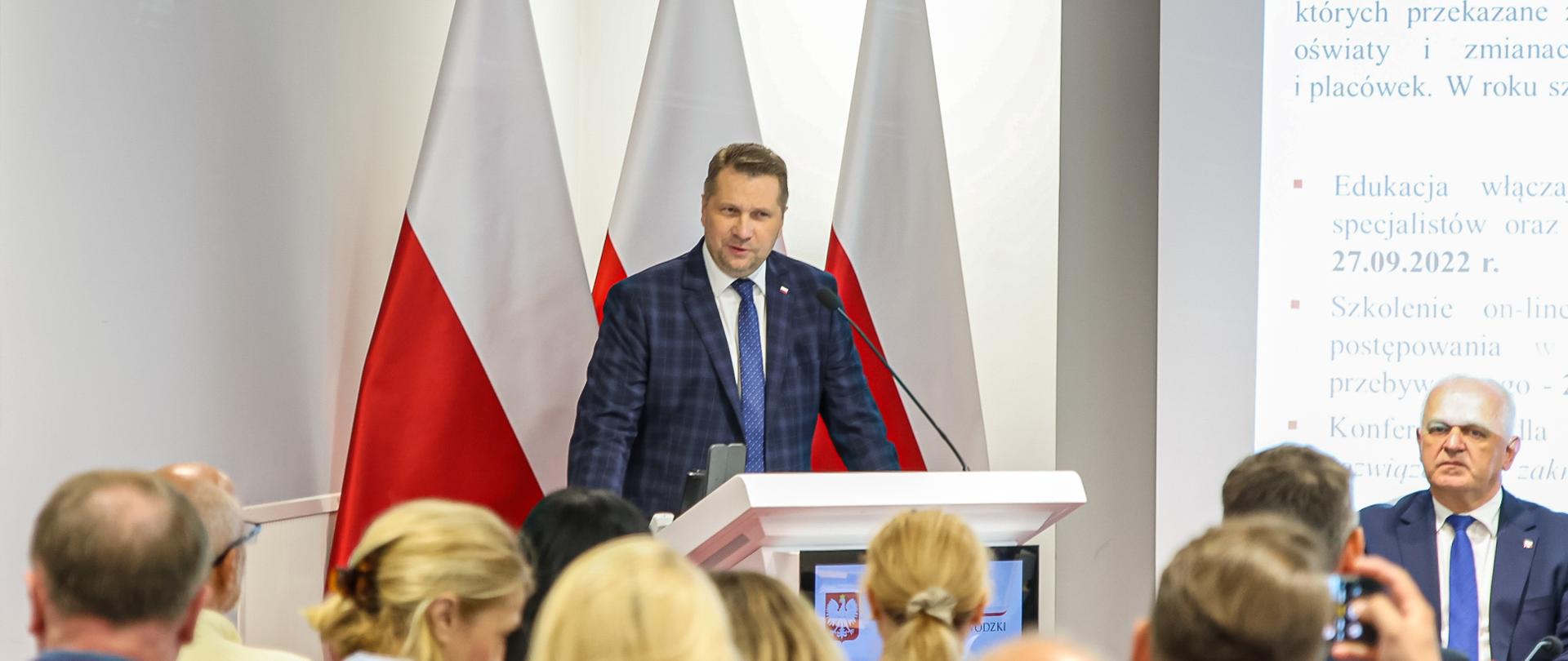 Minister Czarnek przy mównicy, za nim falgi biało-czerwone, na pierwszym planie duża grupa osób siedzących plecami do obiektywu 