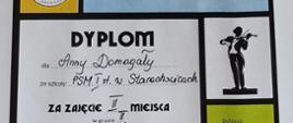 Zdjęcie przedstawia fragment dyplomu. W jego centralnej części widnieje napis:
*DYPlOM*
dla *Anny Domagały*
ze szkoły: *PSM I st. w Starachowicach*
za zajęcie *II miejsca*
w grupie *II*
Po prawej stronie widnieje grafika przedstawiająca sylwetkę osoby grającej na skrzypcach. Niżej, w zielonym polu, znajduje się napis „Bełżyce”. Całość ma charakter oficjalnego dokumentu potwierdzającego udział i osiągnięcie w konkursie muzycznym.