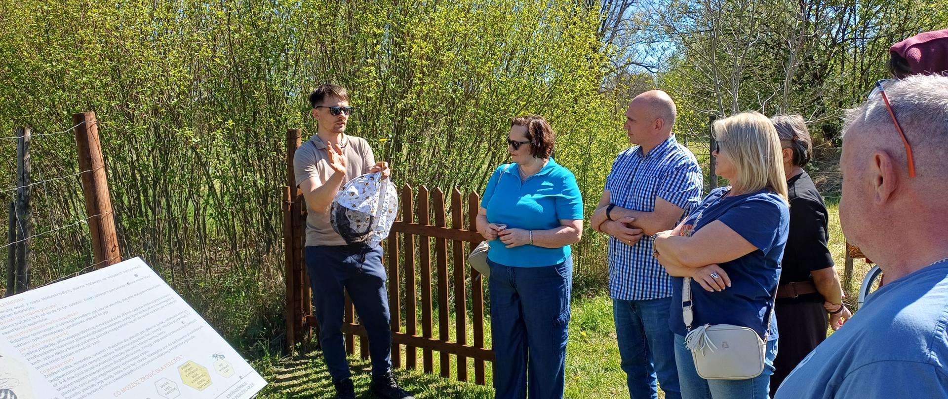 Na zdjęciu znajduje się grupa osób stojących na tle zielonych krzewów i brązowego płotu pośrodku, z lewej strony tablica informacyjna