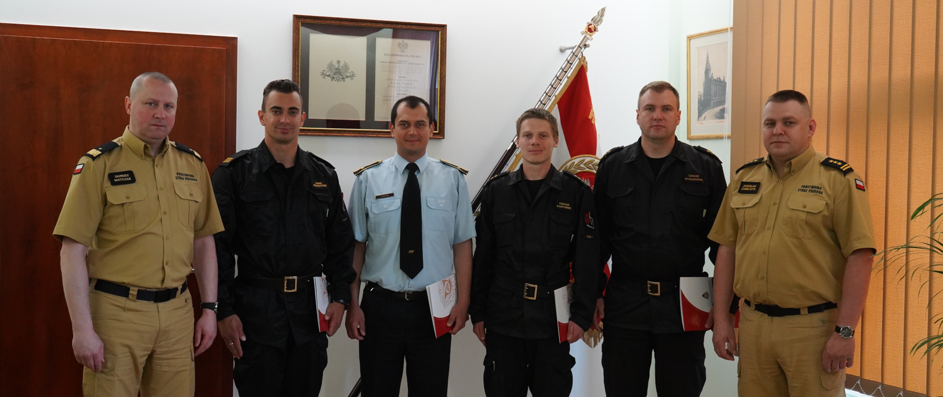 strażacy stoją obok siebie, dwóch z nich na bokach stoi w jasnych mundutrach , jeden w środku stoi w niebieskiej koszulii z krawatem, pozostałych trzech w czranych mundurach , w tle sztandar
