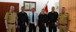 strażacy stoją obok siebie, dwóch z nich na bokach stoi w jasnych mundutrach , jeden w środku stoi w niebieskiej koszulii z krawatem, pozostałych trzech w czranych mundurach , w tle sztandar