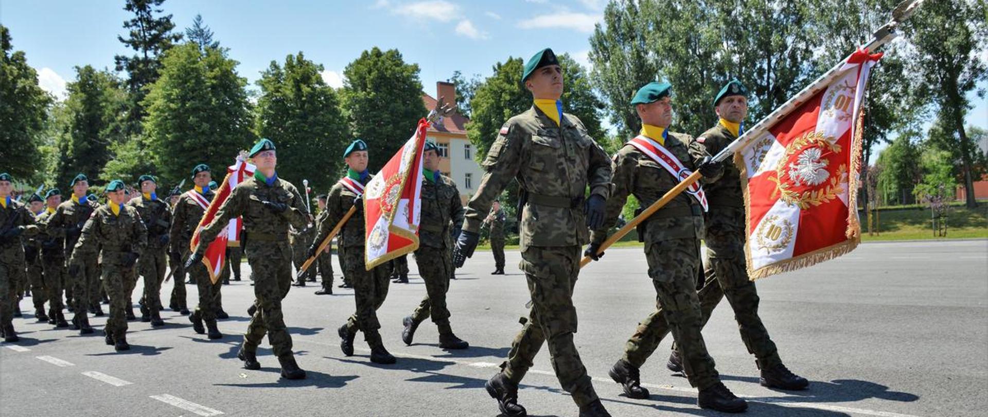 Na zdjęciu szpaler żołnierzy 10 BLOG. W dłoniach trzymają sztandary wojskowe, ubrani w wojskowe mundury. Idą grupami 3 osobowymi. W tle widać drzewa a za drzewami budynek jednostki wojskowej. 