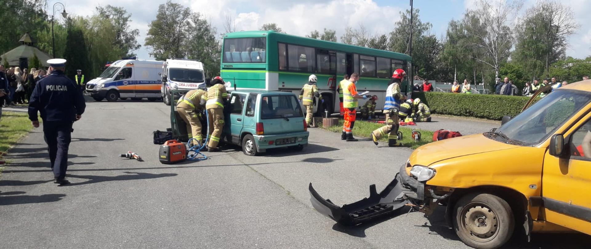 Na zdjęciu wypadek drogowy z udziałem autobusu oraz dwóch samochodów osobowych. Z prawej strony kadru widzimy przód samochodu osobowego, koloru żółtego, urwany zderzak koloru czarnego leży na asfalcie. Środek kadru to tył zielonego samochodu osobowego biorącego udział w wypadku. Przy samochodzie stoi dwóch strażaków, którzy zaglądają do środka przez otwarte drzwi. Strażacy ubrani w ubranie specjalne koloru piaskowego, na głowach czerwone hełmy. Dalej za samochodem osobowym widzimy zielony autobus przy, którym również znajdują się pochyleni strażacy i udzielają pomocy osobie poszkodowanej. Lewa strona kadru to dwie białe karetki oraz grupa ludzi przyglądających się całek akcji.