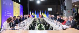 Kobiety i mężczyźni siedzą wokół okrągłego stołu. Wokół nich ścianki i flagi Polski, Szwecji, Ukrainy i Unii Europejskiej.