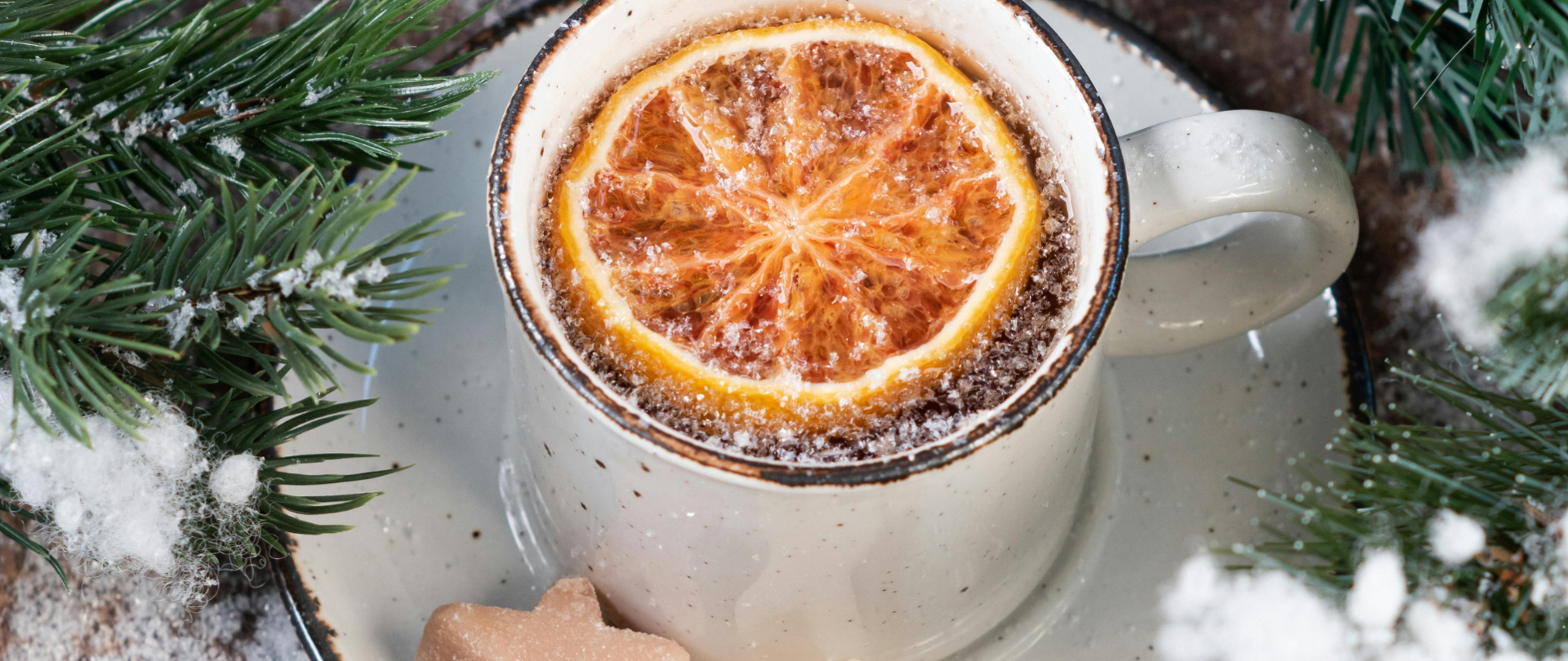 Grafika przedstawia kubek z zimową herbatą, w którym znajduje się plaster pomarańczy, wszystko w zimowej scenerii, otoczone zielonymi gałązkami świerku i przyprószone śniegiem. Na talerzyku obok kubka dwa ciasteczka w kształcie gwiazdki.