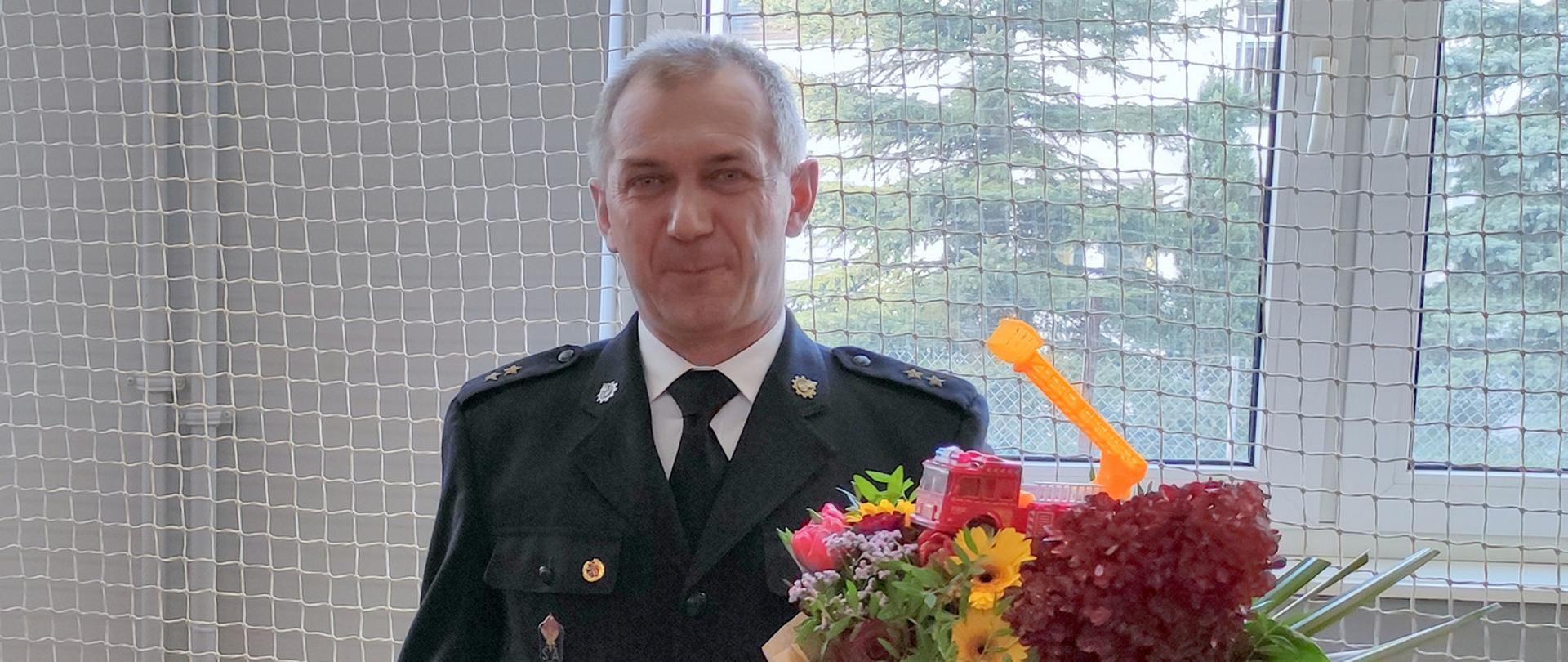 Oficer Państwowej Straży Pożarnej w mundurze wyjściowym z kwiatami i złotym toporkiem z okazji przejścia na emeryturę.