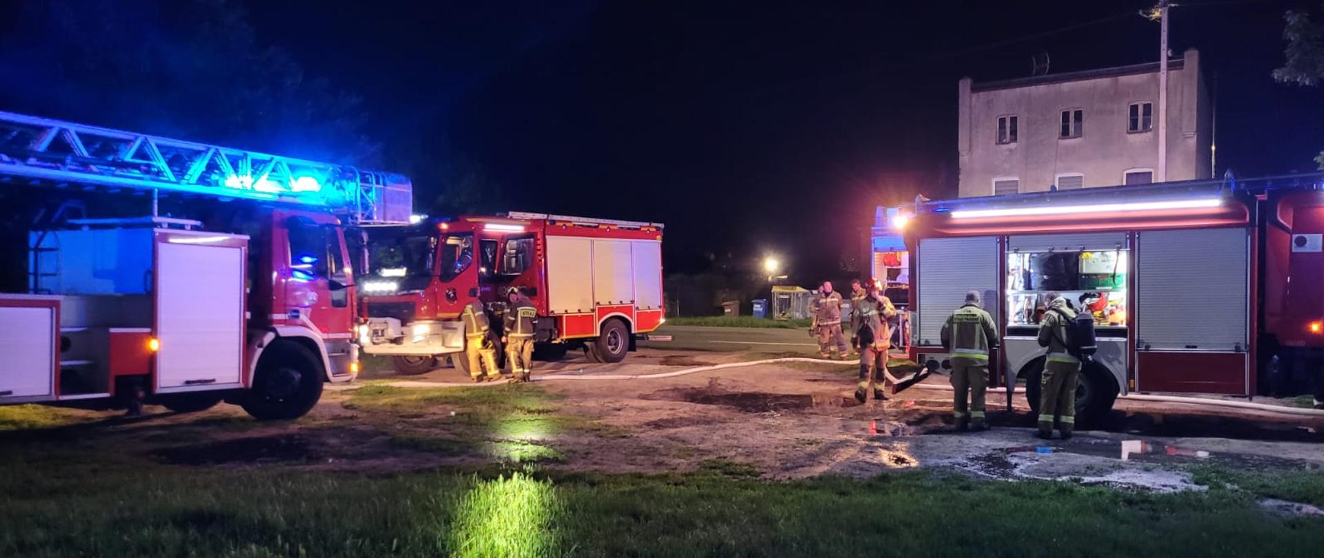 Fotografia przedstawia wozy strażackie i strażaków przy działaniach gaśniczych w porze nocnej.