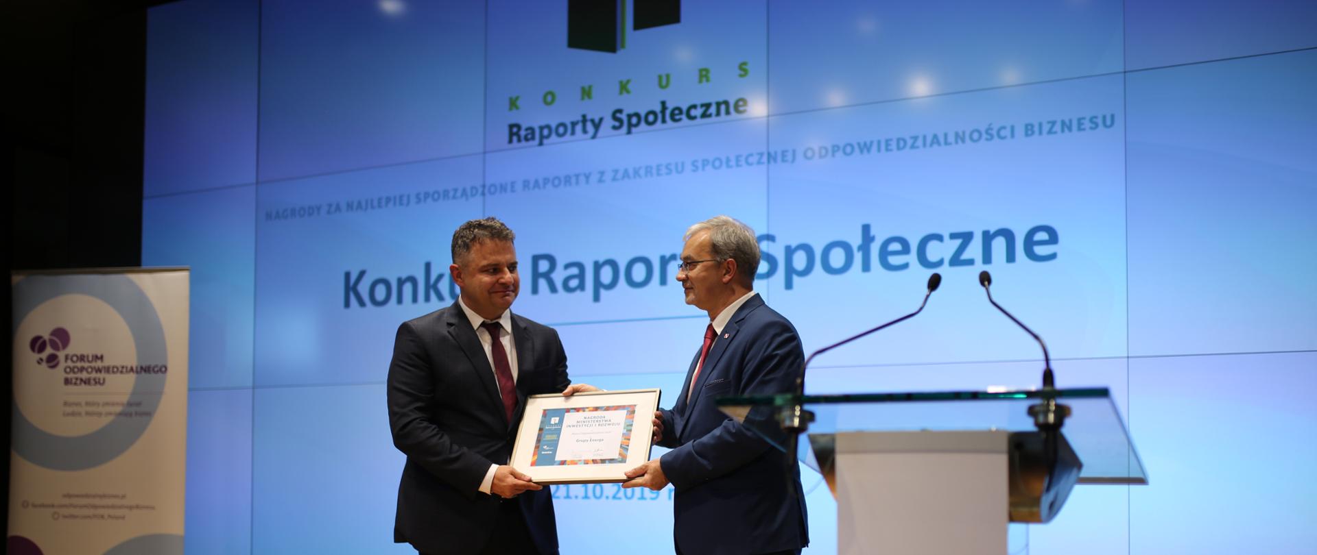 Minister Jerzy Kwieciński wręcza dyplom przedstawicielowi formy Enea. W tle wyświetlony jest napis "Konkurs Raporty Społeczne"