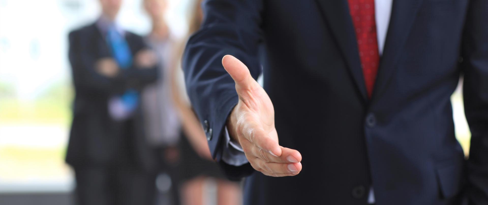 Wyciągnięta dłoń mężczyzny w garniturze a w tle trzy rozmyte postacie