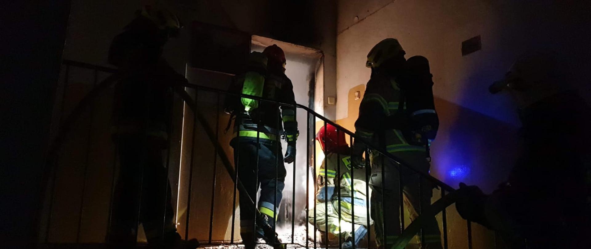 Obraz przedstawia strażaków na klatce schodowej przed otwartym wejściem do mieszkania z widocznym zadymieniem. Klatka schodowa oświetlona tylko latarkami, strażacy w ubraniach specjalnych z aparatami ochrony dróg oddechowych. Jeden ze strażaków kuca przed mieszkaniem obserwując wnętrze mieszkania.