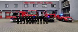 Zdjęcie przedstawia strażaków stojących przed garażem podczas oddawania hołdu zmarłemu strażakowi. Strażacy ubrani w ubrania koszarowe. Za nimi stoją samochody strażackie z włączonymi sygnałami świetlnymi. 