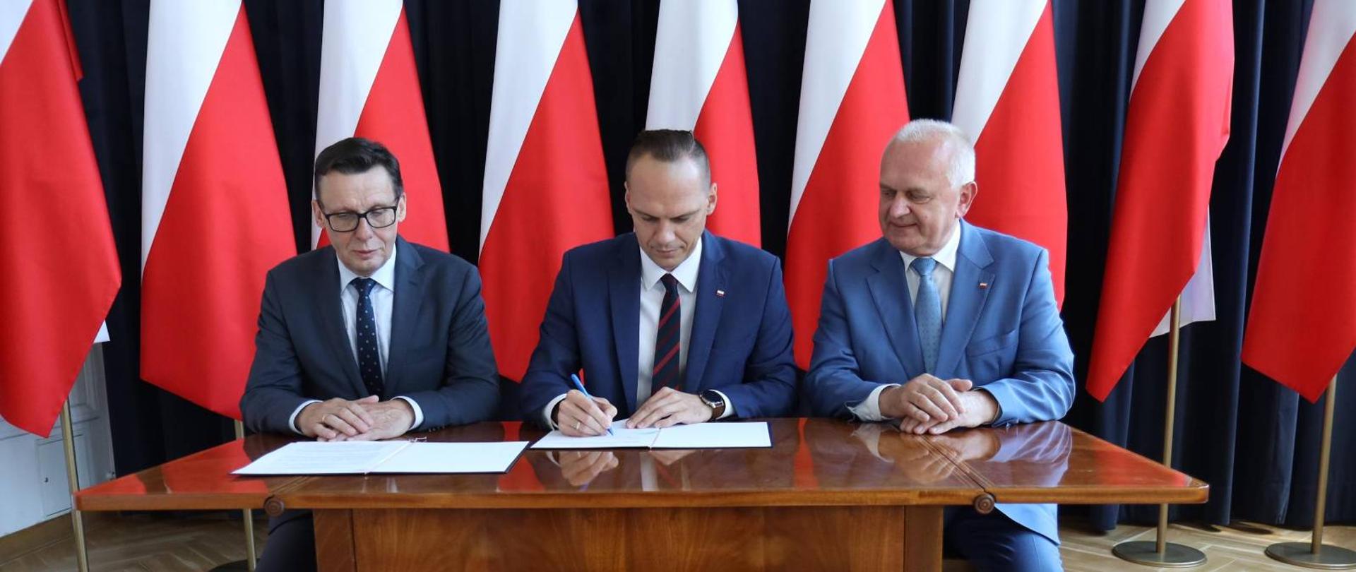 Minister Rafał Weber siedzi przy stole i podpsuje umowę, po lewej stronie siedzi poseł Marek Ast, po prawej stronie wojewoda Władysław Dajczak. W tle biało-czerwone flagi.