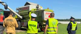 Działania prewencyjne podczas prac polowych. Strażak, policjanci oraz pracownicy Inspekcji Pracy stoją na łące, a przed nimi kombajn.