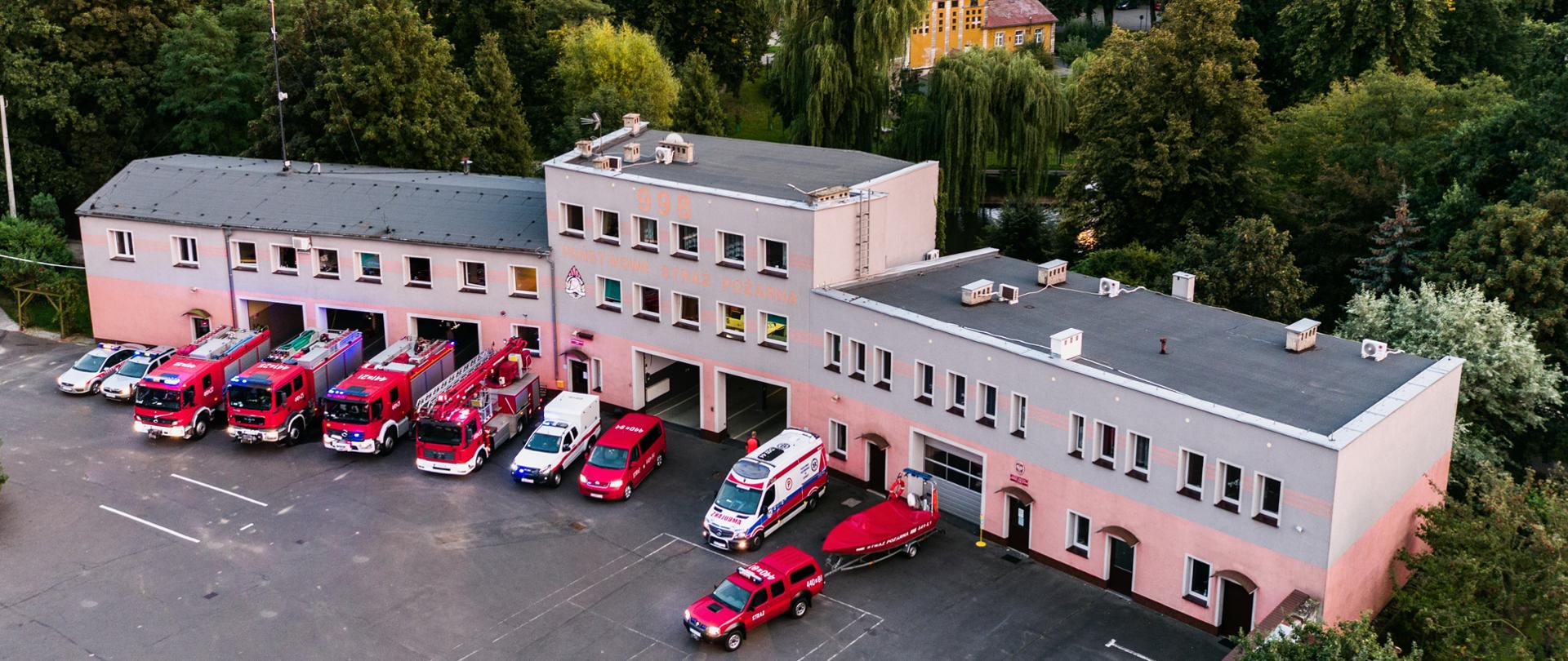 Obraz przedstawia widok na budynek Komendy Powiatowej Państwowej Straży Pożarnej w Jaworze. Przed budynkiem ustawione wszystkie pojazdy pożarnicze komendy oraz karetka Pogotowia Ratunkowego. W tle jaworski park.