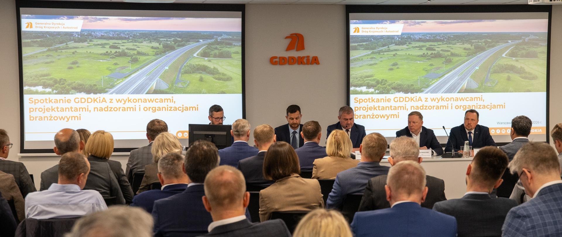 Panel dyskusyjny podczas spotkania GDDKiA z przedstawicielami branży budowlanej; przy stole siedzi kilka osób z mikrofonami i laptopami, a na ekranie za nimi widoczna jest prezentacja z tytułem dotyczącym współpracy przy realizacji inwestycji oraz data w Warszawie.