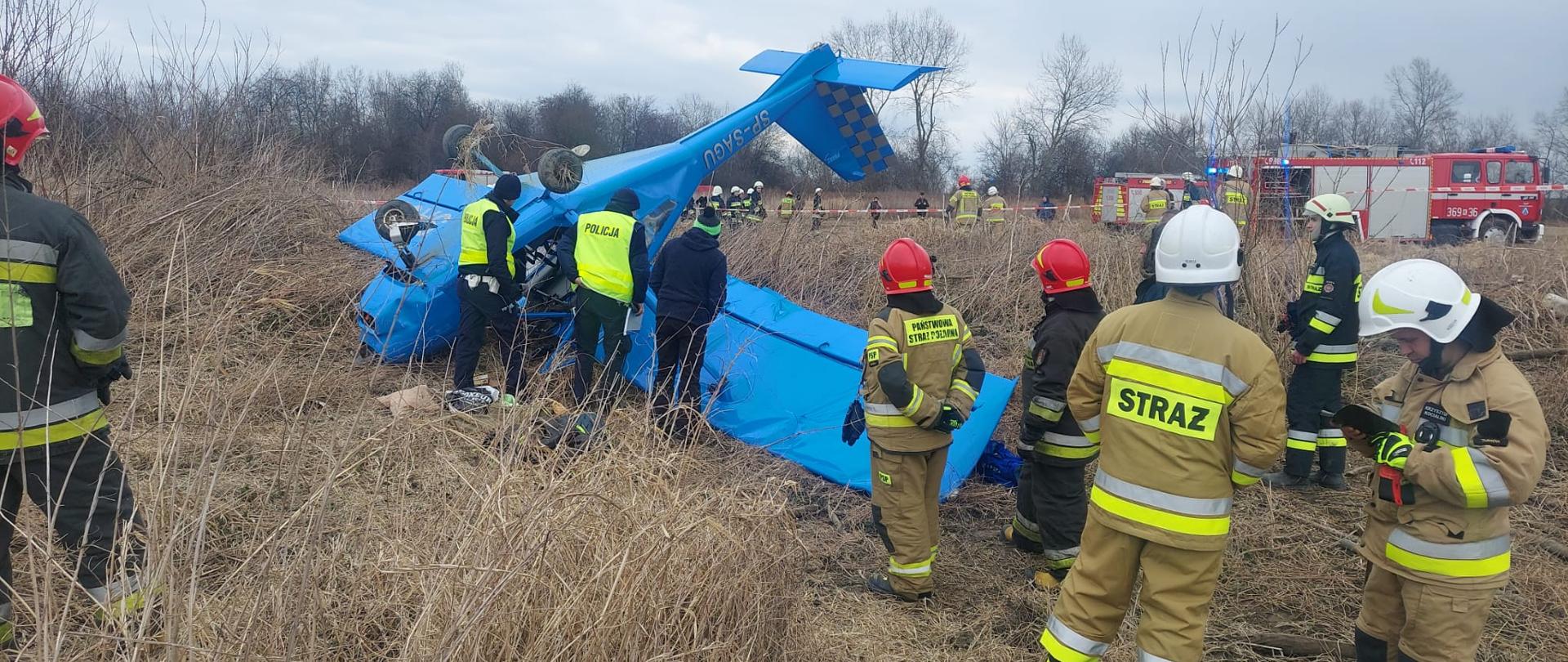 Na zdjęciu widoczna awionetka koloru niebieskiego leżąca na łące do góry kołami. Obok samolotu znajduje się funkcjonariusz Policji sprawdzający wnętrze awionetki. W tle widoczne są wozy bojowe strażaków i strażacy.