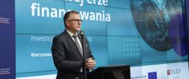 Prezes Polskiej Agencji Rozwoju Przedsiębiorczości Krzysztof Gulda stoi przy mównicy i przemawia, za jego plecami telebim informujący o nowej formie finansowania firm