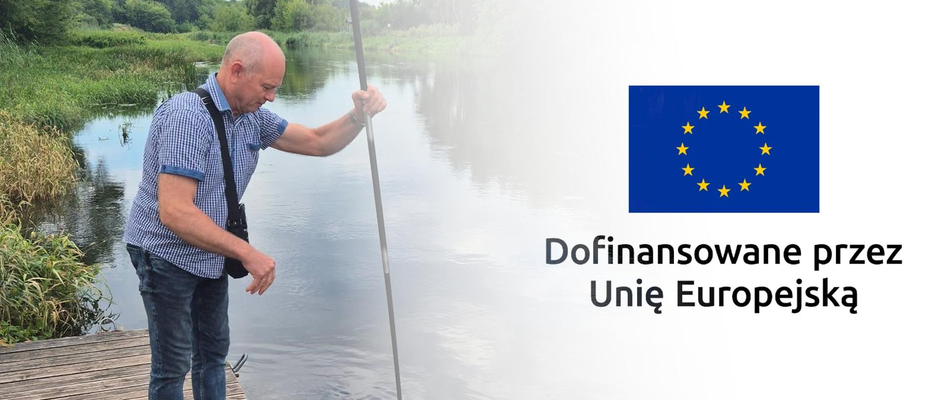 Mężczyzna stojący na pomoście pobiera próbkę z rzeki za pomocą długiego drążka; obok widnieje flaga Unii Europejskiej i napis „Dofinansowane przez Unię Europejską”.