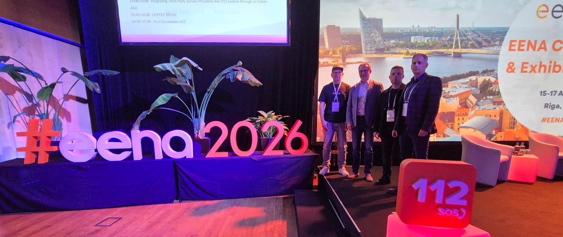 Uczestnicy konferencji EENA 2026 stoją na scenie obok napisu „#eena2026” i podświetlanego symbolu „112”, w tle ekran z widokiem Rygi.