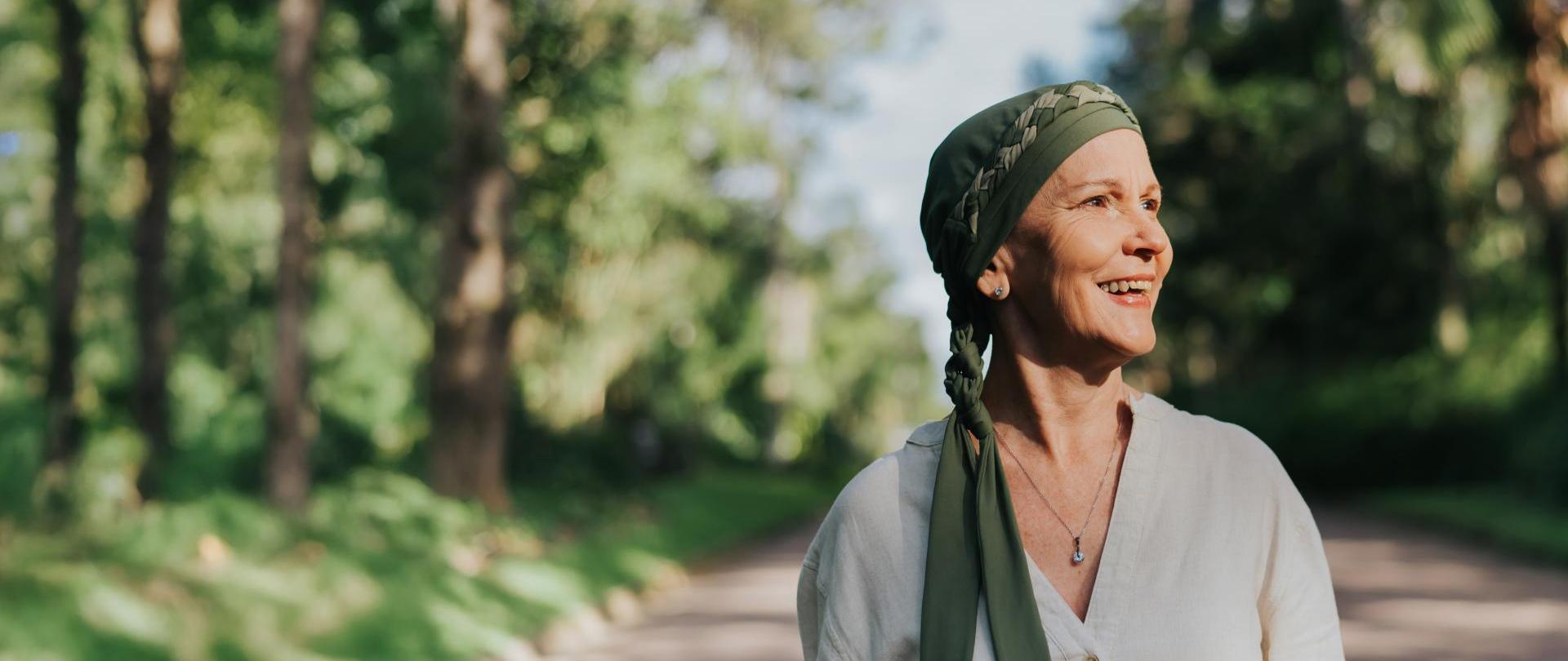 Starsza kobieta w jasnej bluzce i w oliwkowej chuście na głowie z subtelnym uśmiechem, która spaceruje alejką w parku miejskim. 