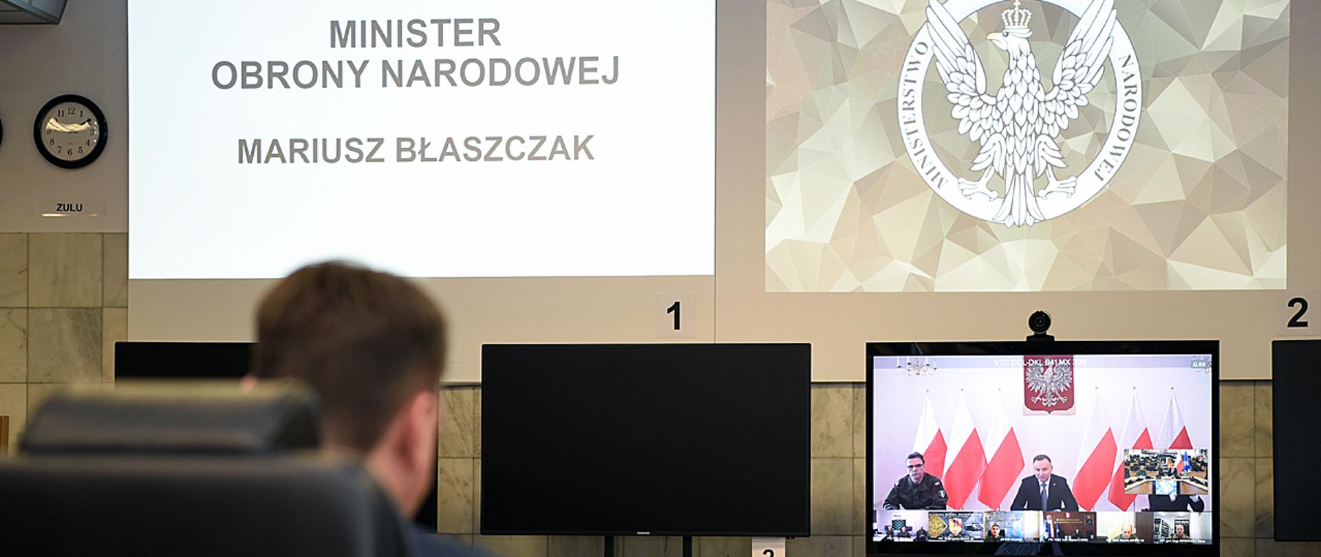 Szef MON podczas wideokonferencji na dużej sali. Na monitorze Prezydent Andrzej Duda i dowódcy Wojska Polskiego. Na ścianie logo MON i napis "MINISTER OBRONY NARODOWEJ MARIUSZ BŁASZCZAK".