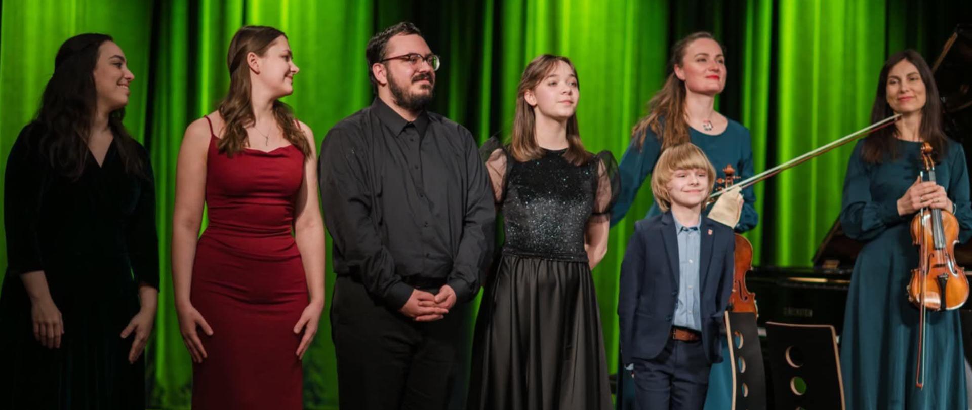 kilkuosobowa grupa dorosłych i jedno dziecko stojący na scenie, po prawej dwa krzesła, w tle zielona kotara