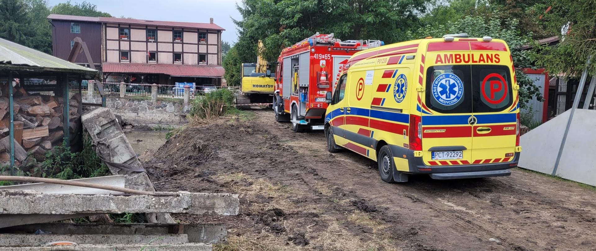 Pojazd pogotowia ratunkowego i straży pożarnej na tle budynku przy rzece Gulczanka oraz koparki.