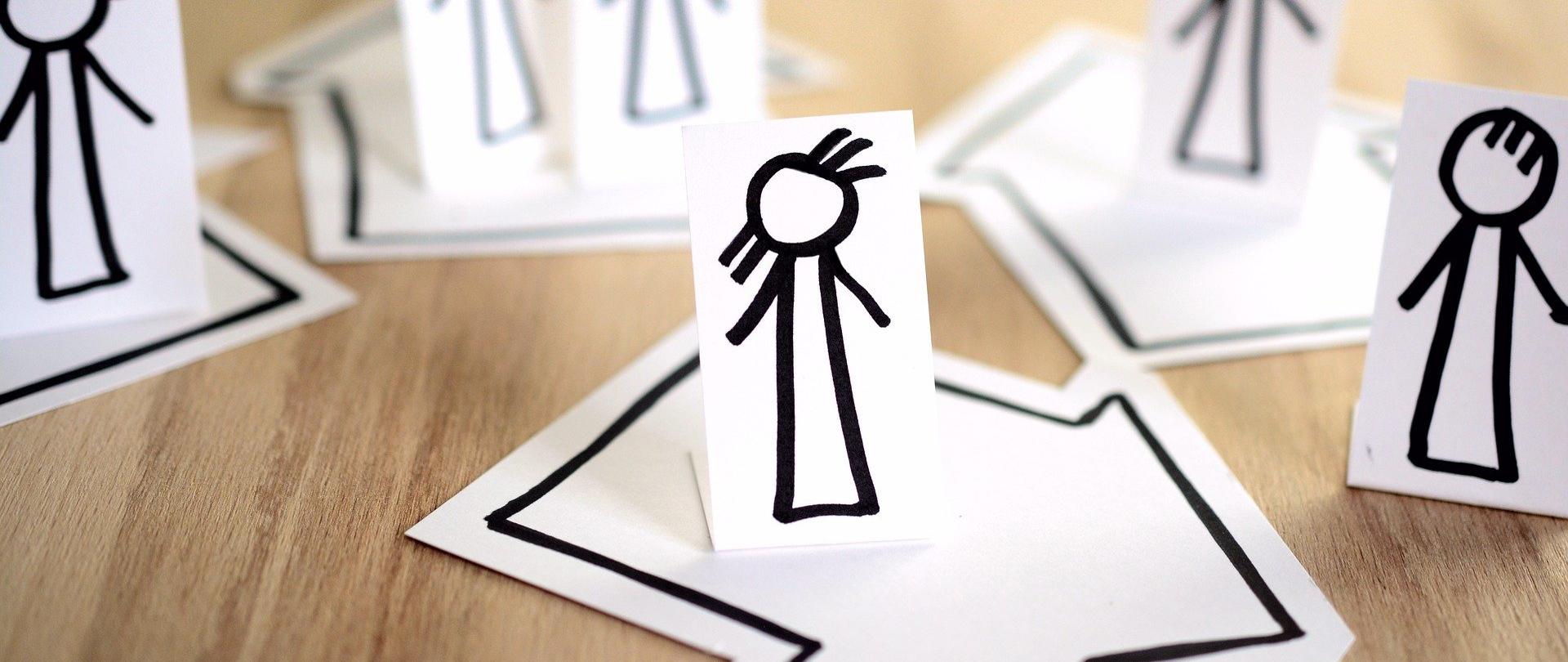 Zdjęcie przedstawia papierowe figury ludzI, którzy stoją na wyciętych także z papieru domkach. Kompozycja ma nawiązywać do kwarantanny. Papierowe elementy ułożone są na drewnianym stole. 