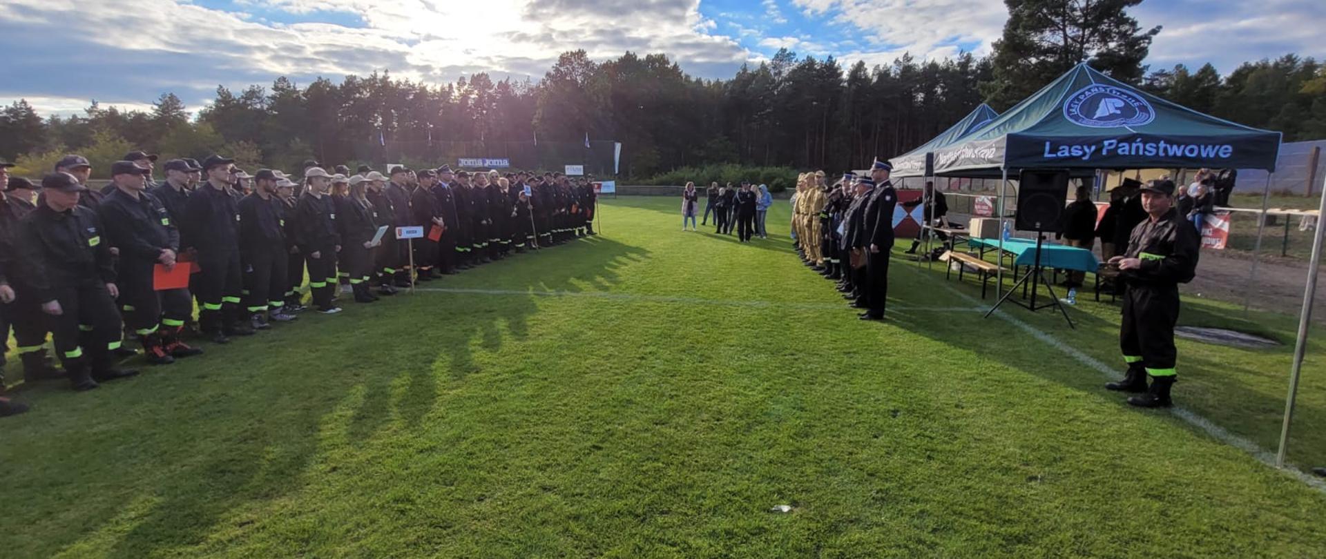 Uroczyste rozpoczęcie zawodów sportowo-pożarniczych OSP, drużyny strażaków ochotników stoją w szeregach naprzeciw sędziów.