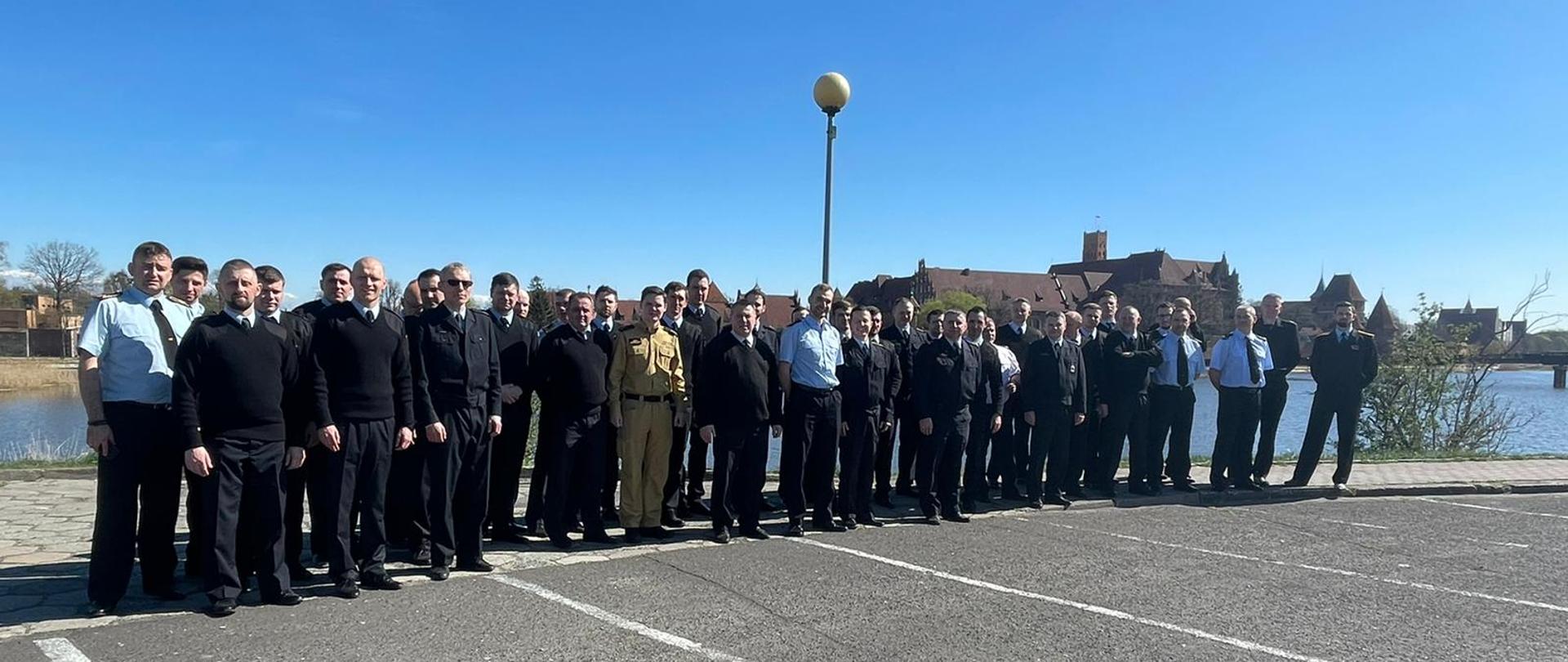 Fotografia przedstawia funkcjonariuszy pionu kontrolno-rozpoznawczego ustawionych na zdjęciu grupowym na tle Zamku Malborskiego oraz rzeki Nogat.