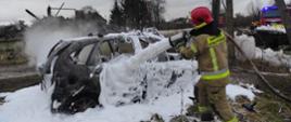 Strażak w piaskowym umundurowaniu i czerwonym hełmie dogasza auto przy pomocy piany. Wrak całkowicie pokryty białą pianą